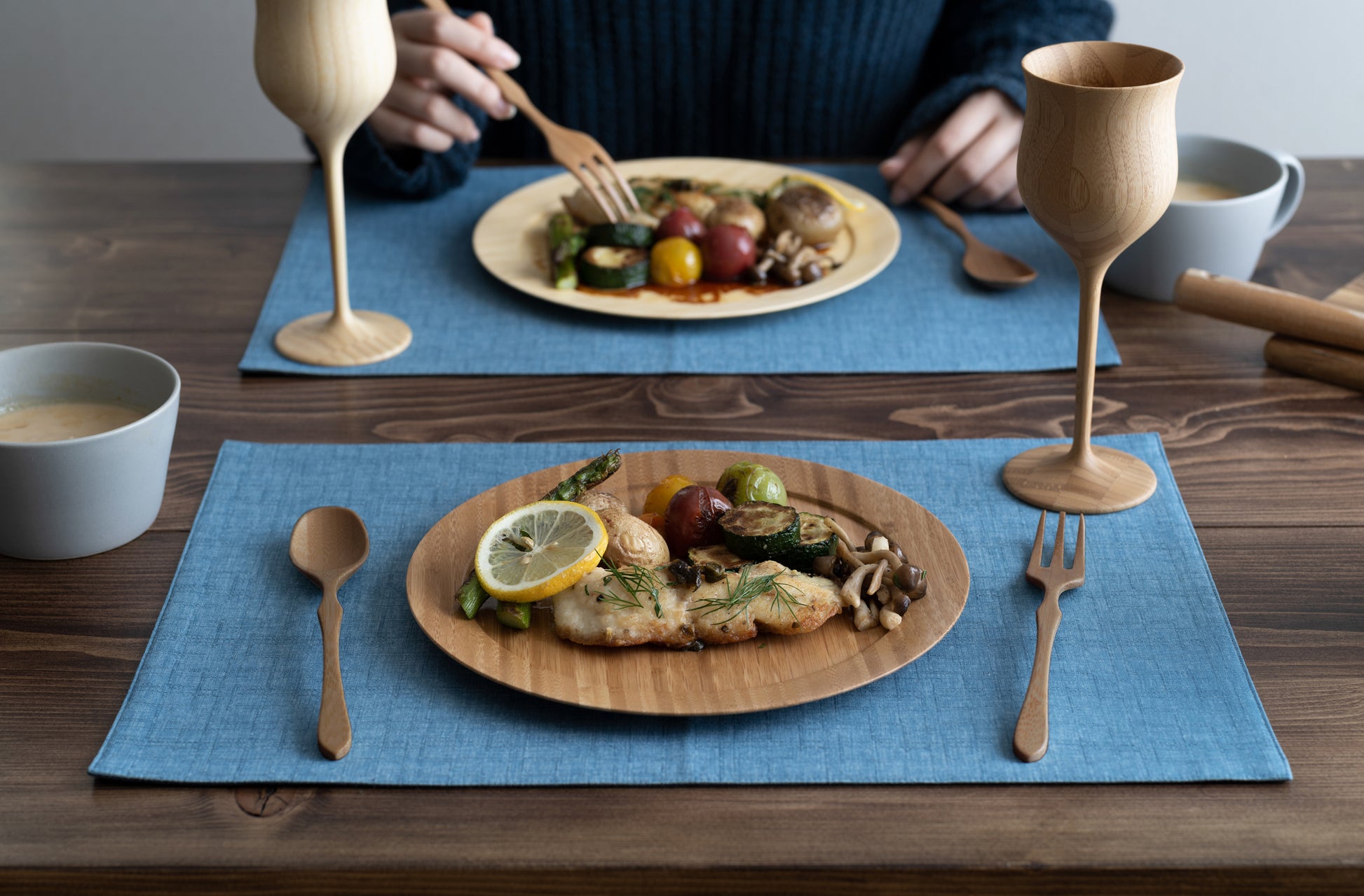 Two eco-friendly round bamboo dinner plates served with grilled fish and roasted vegetables on blue placemats, featuring matching bamboo stemware and wooden cutlery on a rustic table.