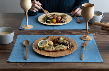 Two eco-friendly round bamboo dinner plates served with grilled fish and roasted vegetables on blue placemats, featuring matching bamboo stemware and wooden cutlery on a rustic table.